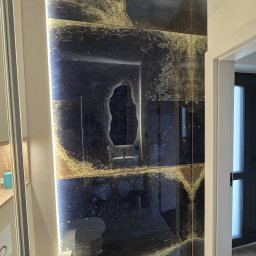 Dawid Gol Usługi Remontowo-Budowlane - Błyszcząca ściana w łazience wyłożona ciemnymi płytkami z efektem złota, odbijająca przestrzeń. Widoczny fragment toalety i drzwi.