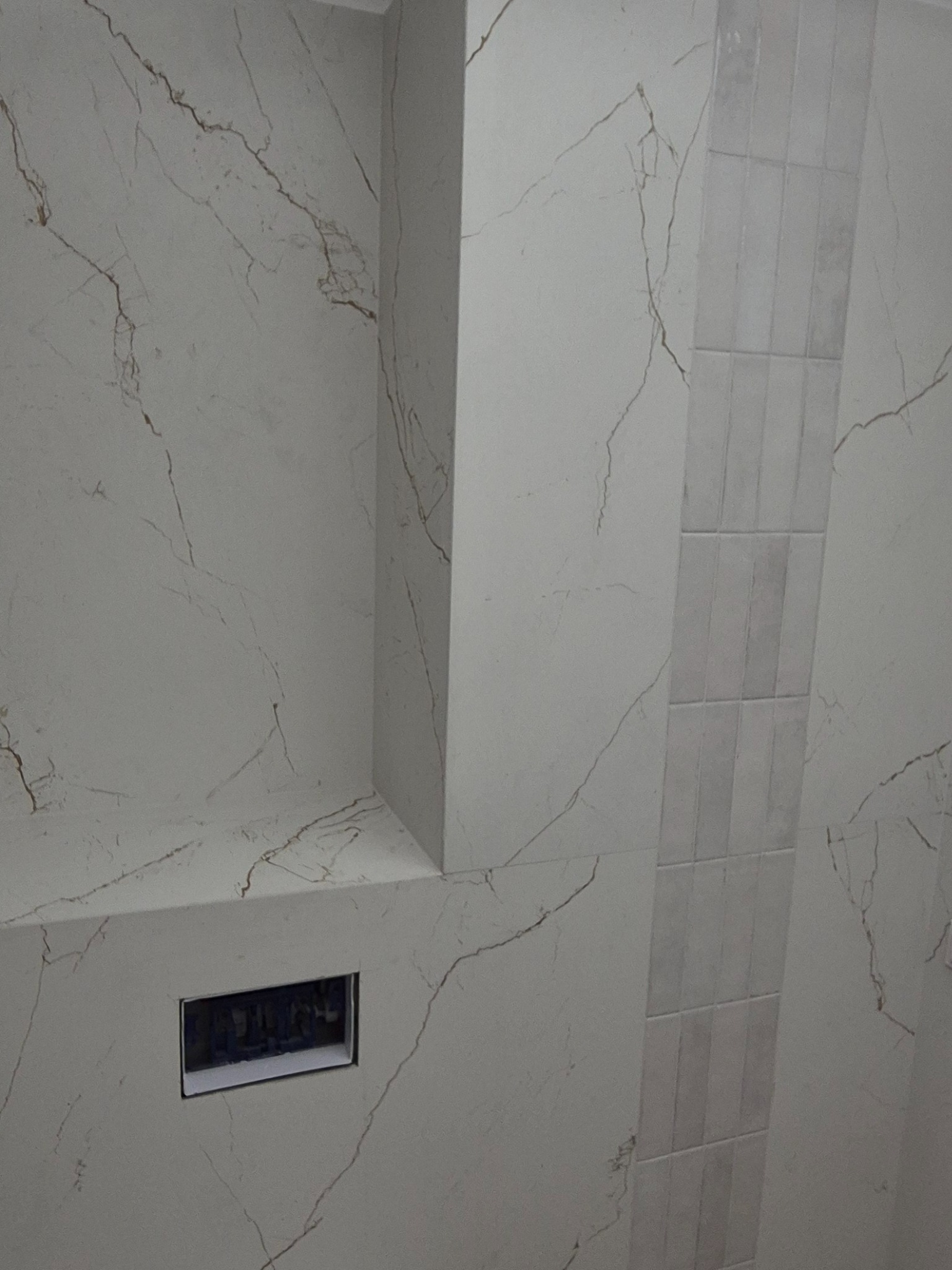 Wykończona ściana w łazience z płytkami imitującymi marmur z delikatnym wzorem żyłkowania i wstawką z płytek w innym odcieniu. Widoczny otwór na gniazdko.