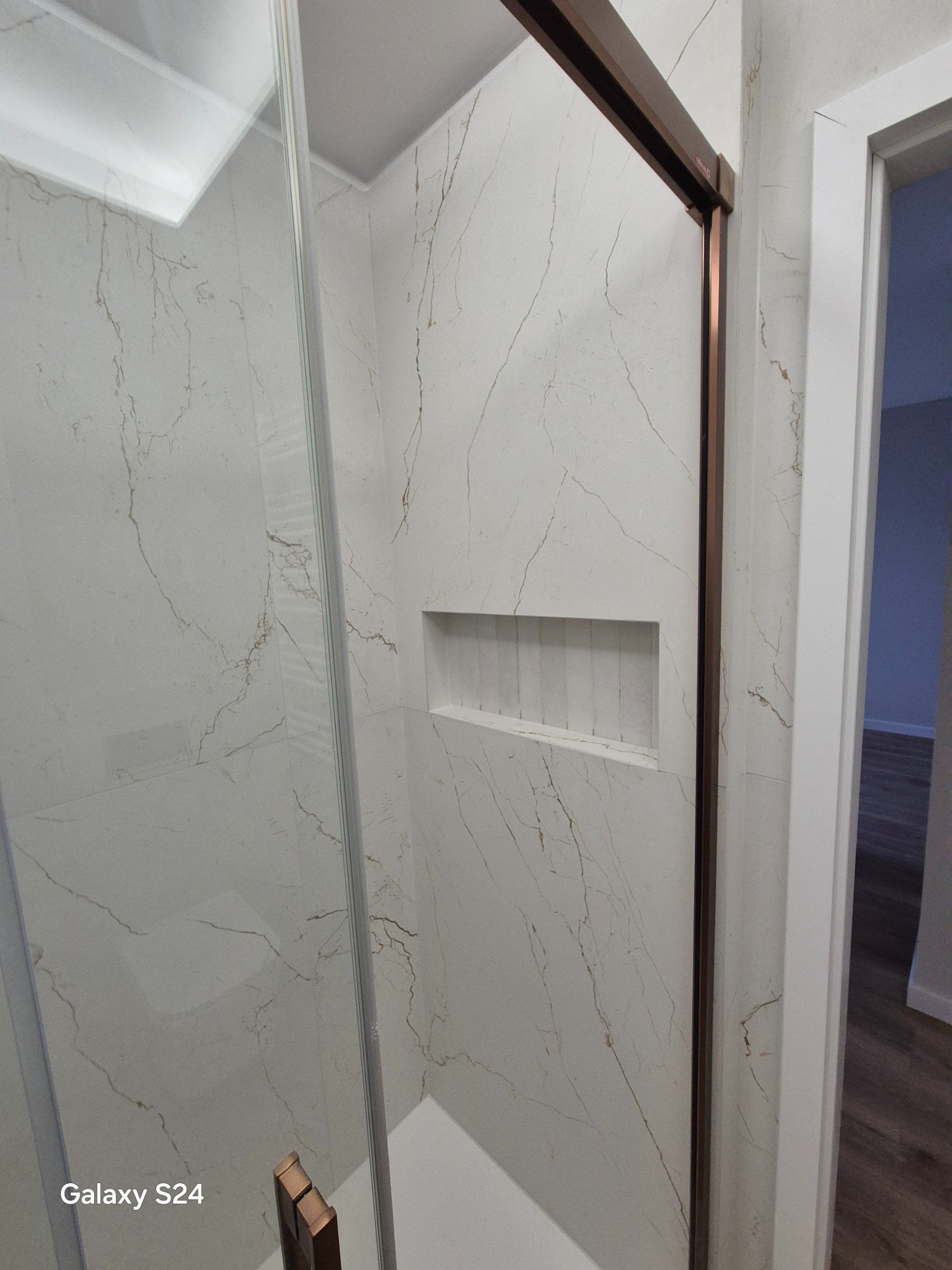 Kabina prysznicowa z białymi płytkami imitującymi marmur, z wnęką na kosmetyki i drzwiami przesuwnymi z brązowymi ramami. Widok na fragment łazienki z podłogą.