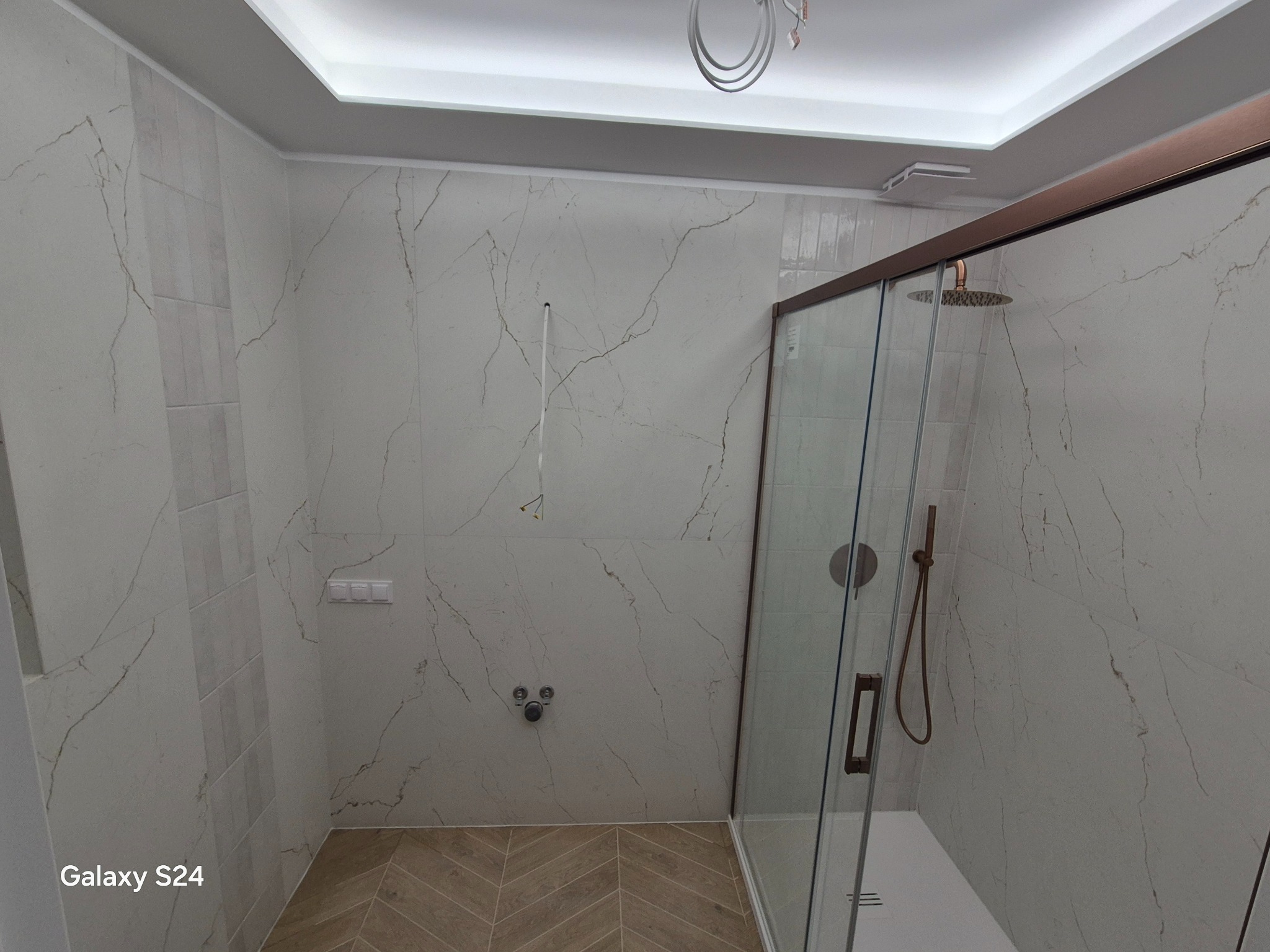 Wykończona łazienka z kabiną prysznicową typu walk-in, marmurowe płytki na ścianach i podłodze ułożonej w jodełkę. Widoczne elementy armatury w kolorze miedzi.