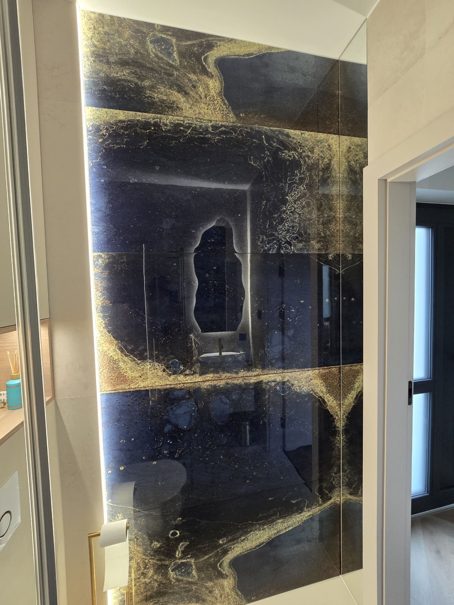 Błyszcząca ściana w łazience wyłożona ciemnymi płytkami z efektem złota, odbijająca przestrzeń. Widoczny fragment toalety i drzwi.