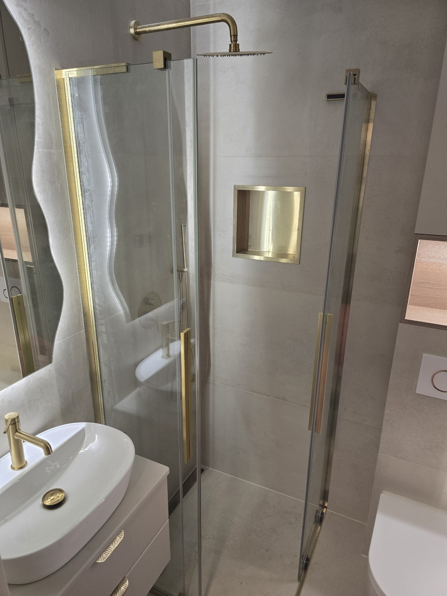 Nowoczesna łazienka z kabiną prysznicową typu walk-in z złotymi detalami, umywalką nablatową i wnęką w ścianie. Minimalistyczny design, jasne płytki, eleganckie wykończenie.