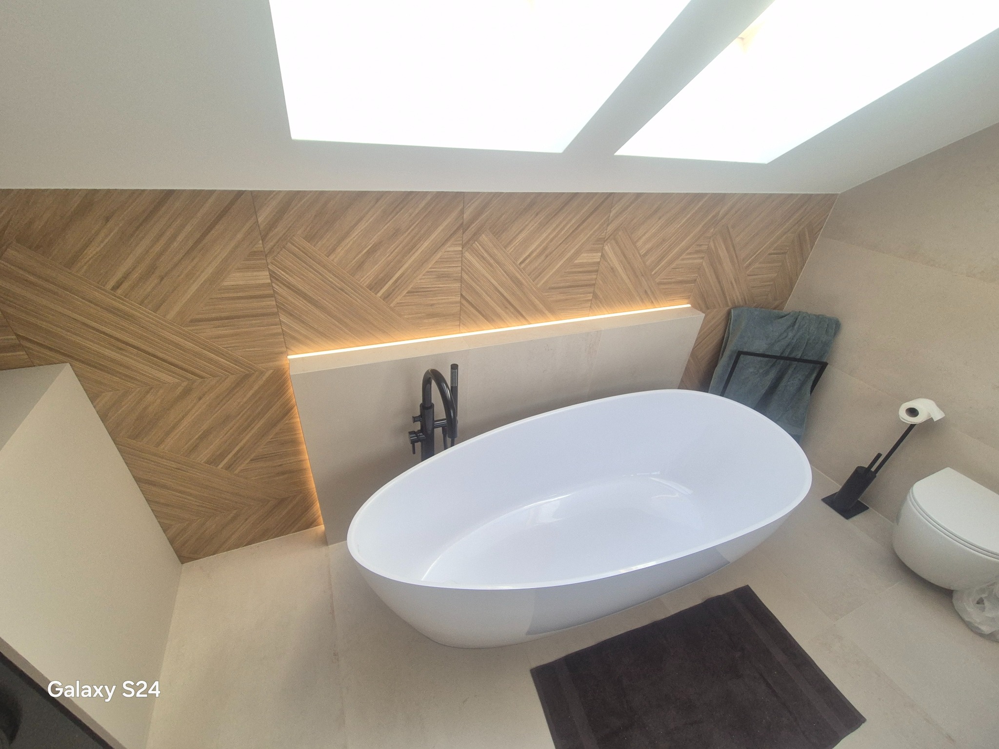 Nowoczesna łazienka z wolnostojącą białą wanną, drewnianą mozaiką na ścianie z podświetleniem LED i oknem w suficie. Minimalistyczny design, jasne płytki podłogowe.