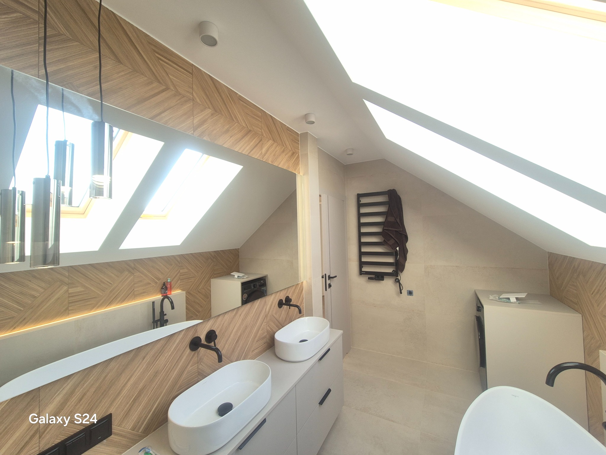 Jasna łazienka na poddaszu z drewnopodobnymi płytkami, dwiema umywalkami nablatowymi i czarną armaturą. Widoczne okno dachowe i grzejnik drabinkowy.