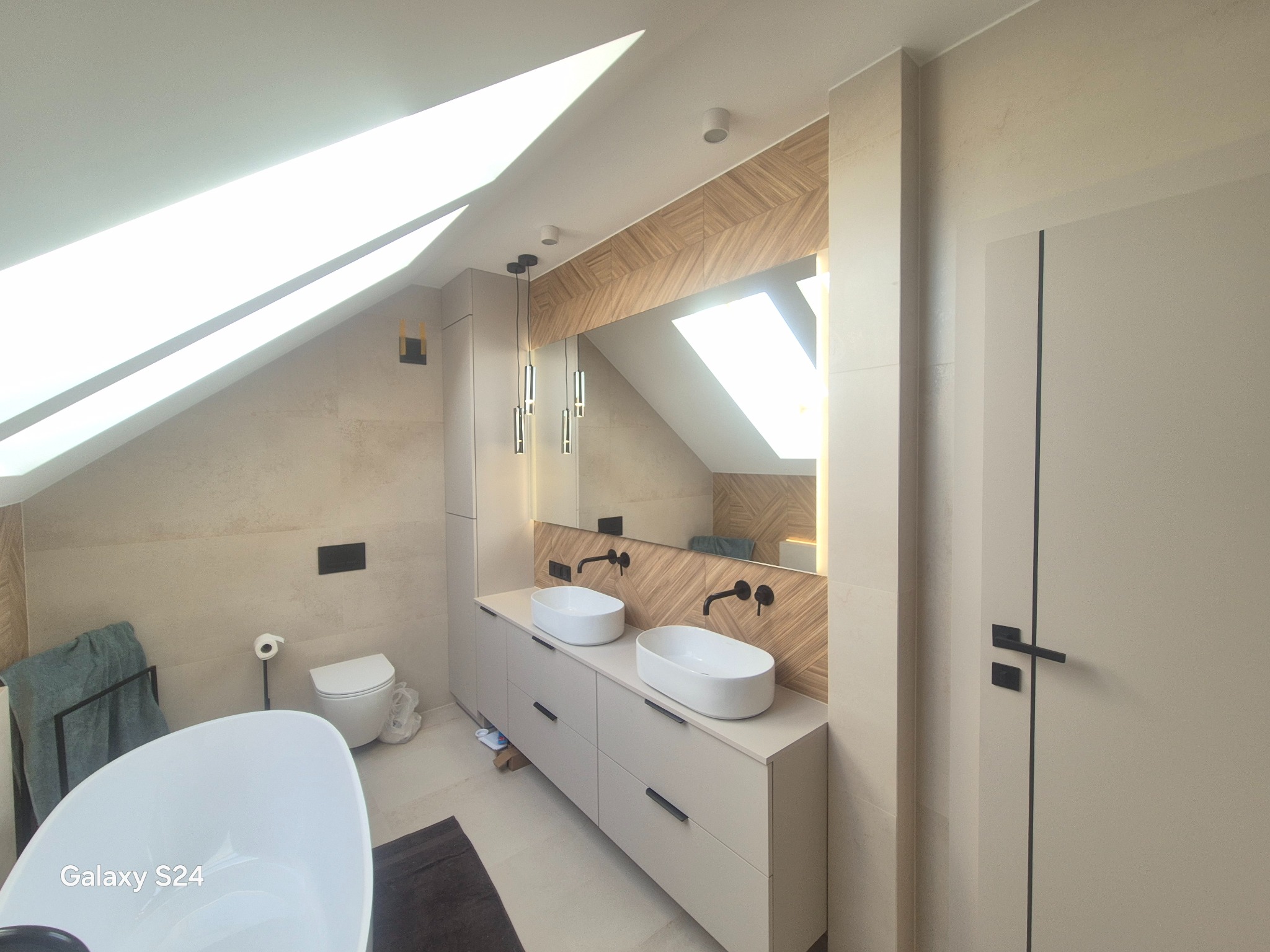 Jasna łazienka na poddaszu z dwoma umywalkami nablatowymi, drewnianą mozaiką nad blatem i oknem dachowym w lustrze. Minimalistyczny design, neutralna kolorystyka.