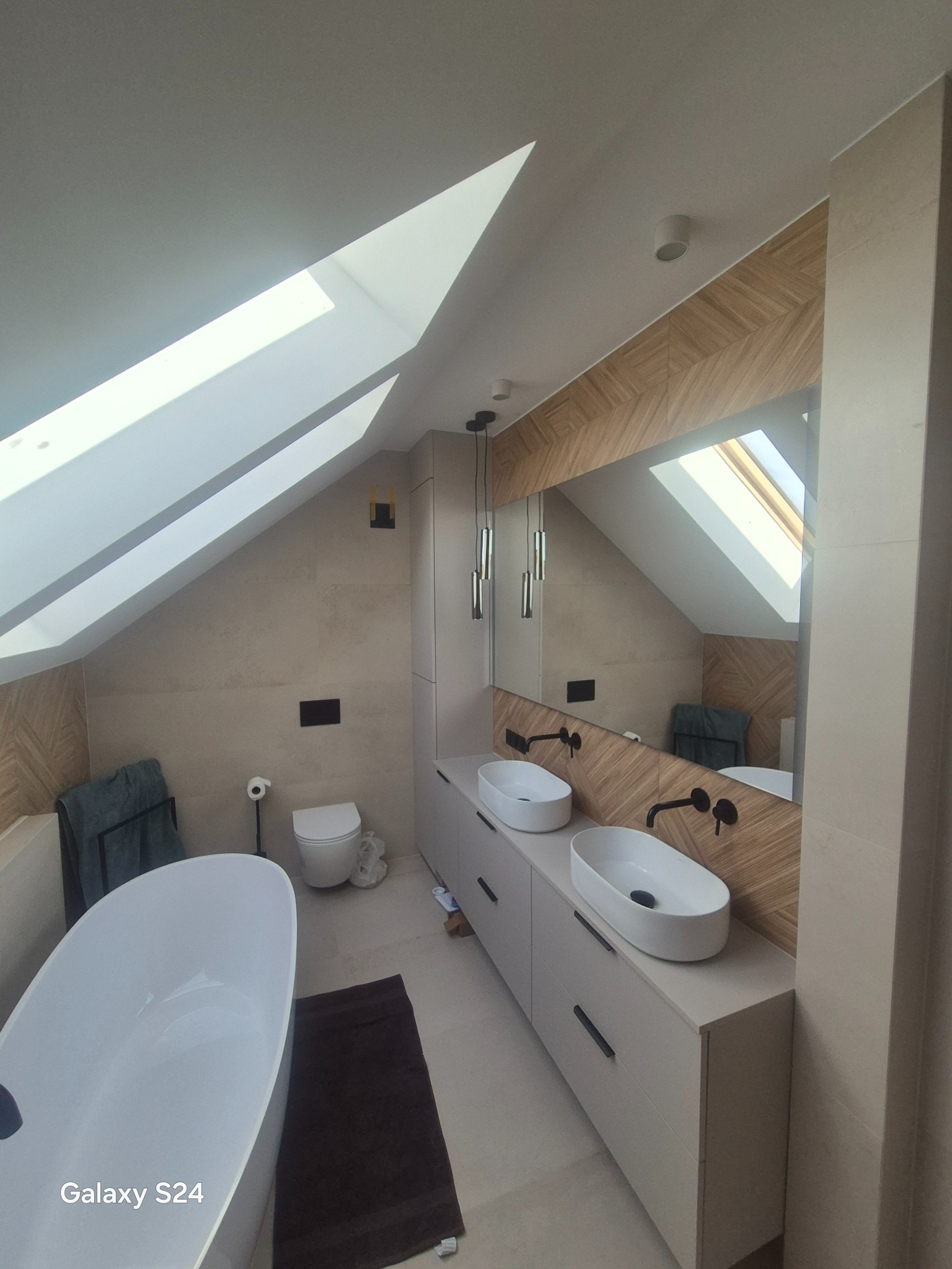 Nowoczesna łazienka na poddaszu z wanną wolnostojącą, dwiema umywalkami nablatowymi i lustrem z drewnianą ramą. Uwagę przykuwają skosy i okna dachowe.