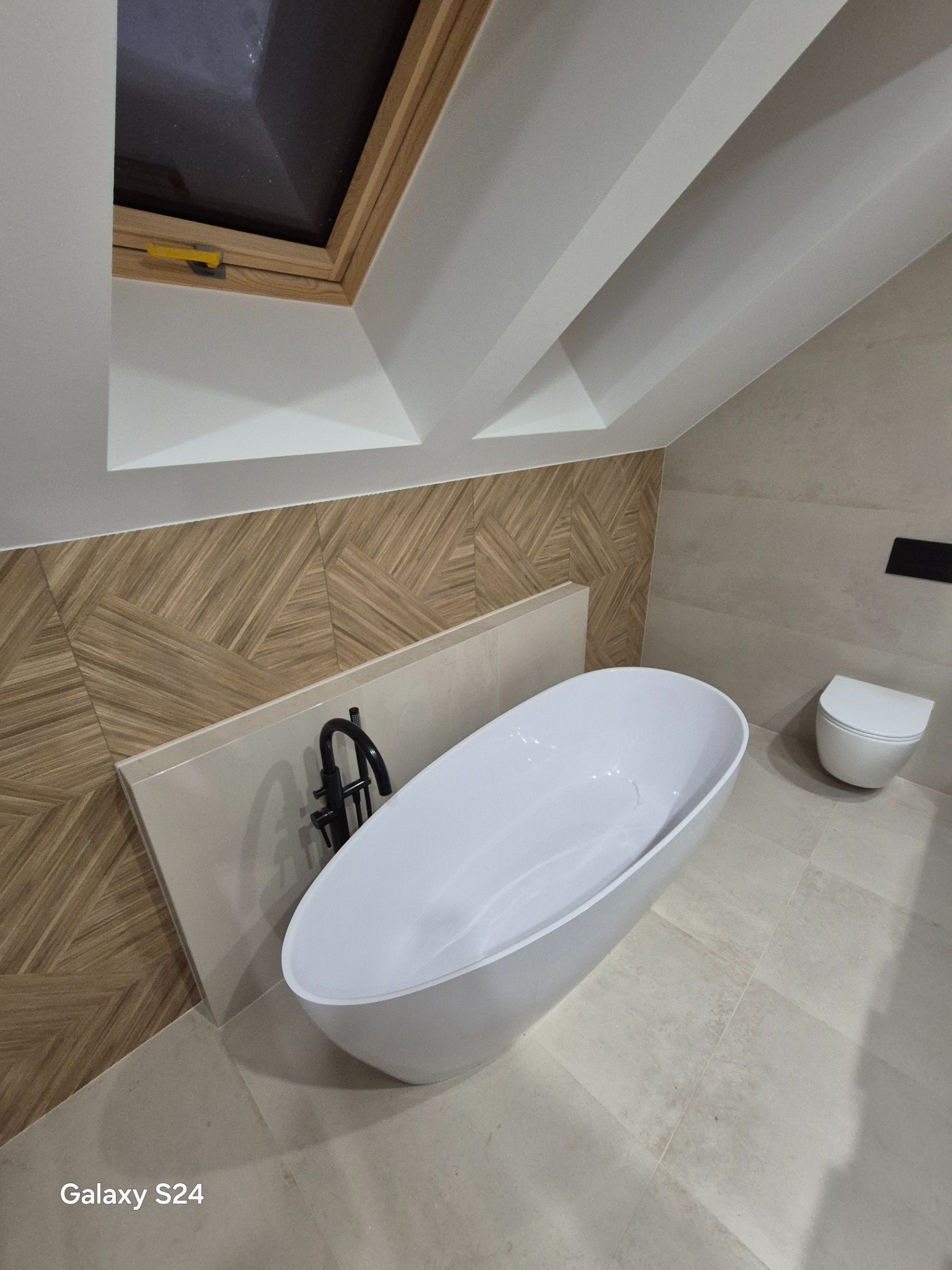 Nowoczesna łazienka na poddaszu z wolnostojącą wanną, drewnopodobnymi płytkami na ścianie i oknem dachowym. Minimalistyczny design i jasna kolorystyka.