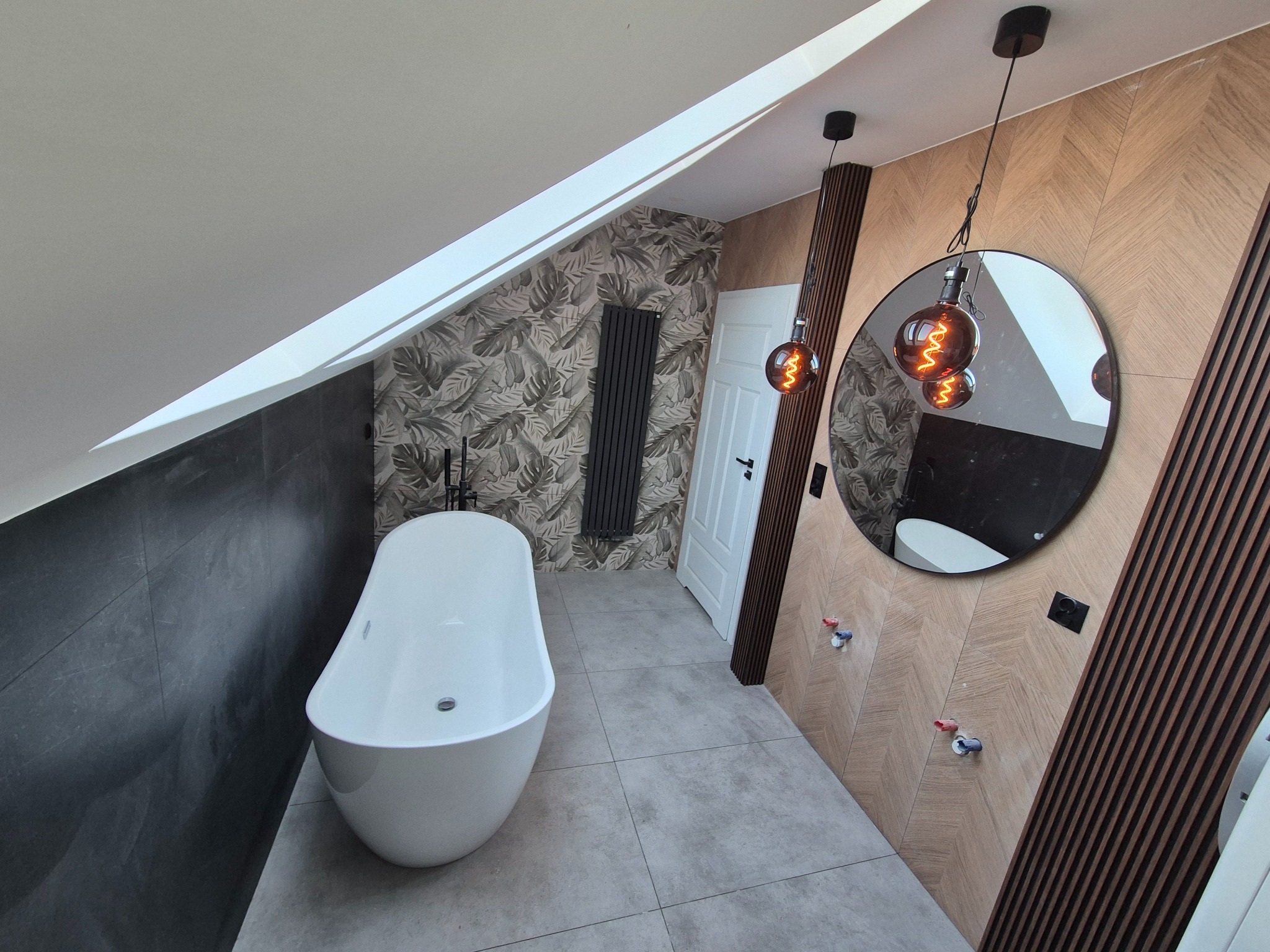 Nowoczesna łazienka na poddaszu z wolnostojącą wanną, szarą armaturą i wzorzystą tapetą. Uwagę przykuwają lampy wiszące z żarówkami Edisona i okrągłe lustro.