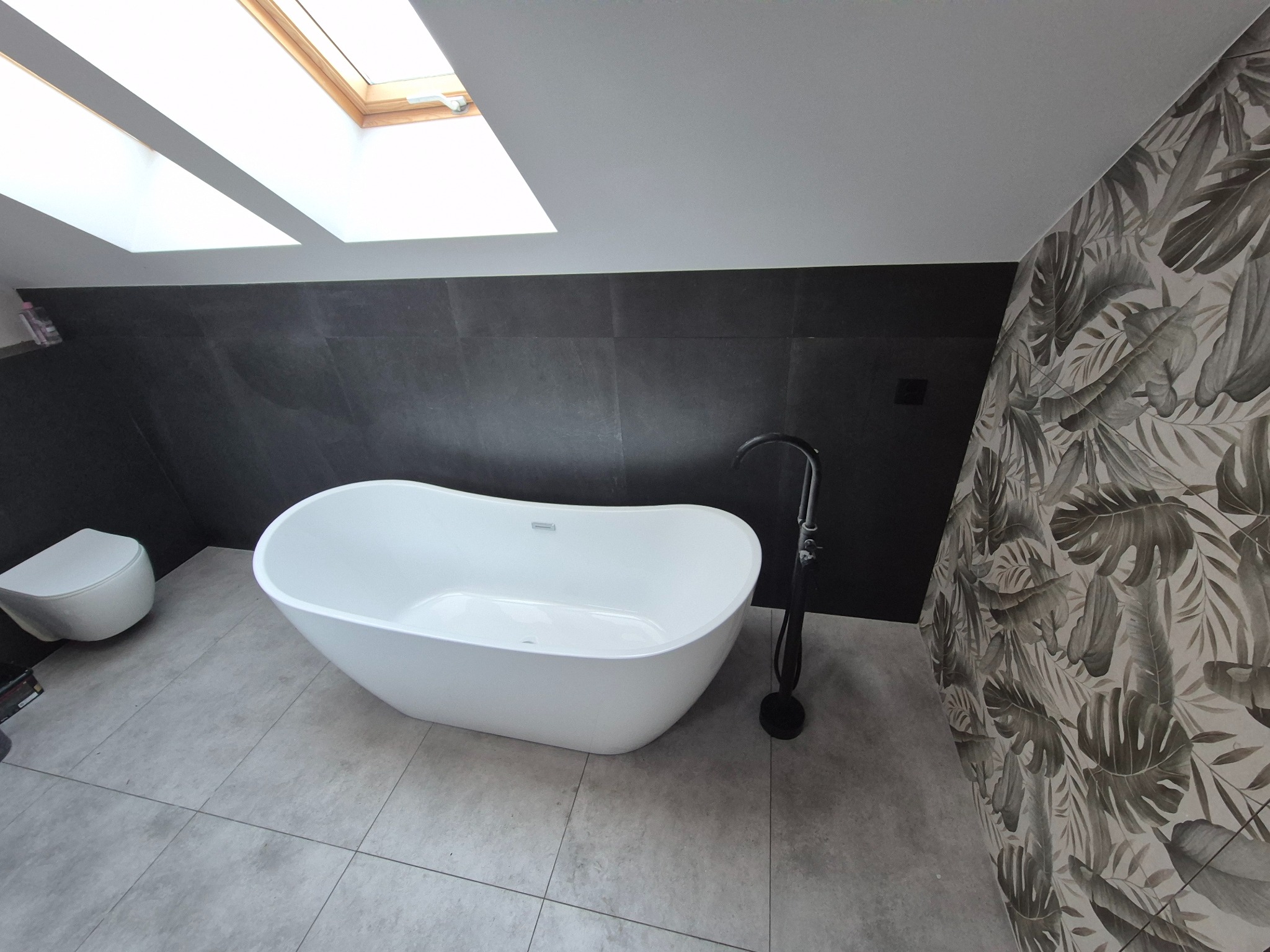 Nowoczesna łazienka na poddaszu z wolnostojącą białą wanną, czarną armaturą i ścianą w tropikalne liście. Okno dachowe doświetla wnętrze.