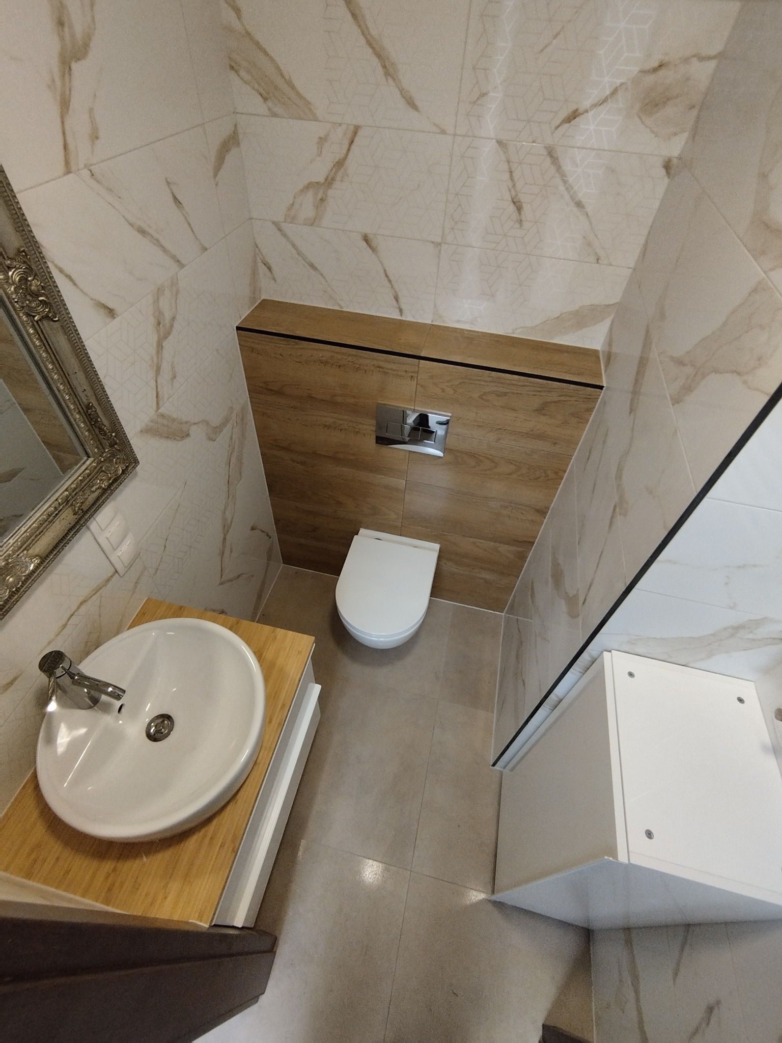 Wykończona łazienka z umywalką na drewnianym blacie, toaletą w zabudowie i marmurowymi płytkami na ścianach. Ujęcie z góry podkreśla przestrzeń i detale.