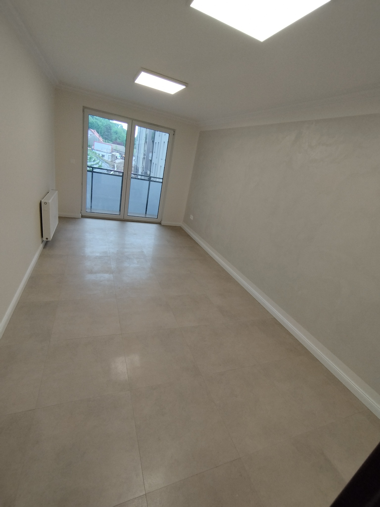 Jasne, wykończone wnętrze pokoju z beżową podłogą z płytek, ścianami w odcieniach bieli i dużym oknem balkonowym z widokiem na zieleń. Minimalistyczny grzejnik przy ścianie.