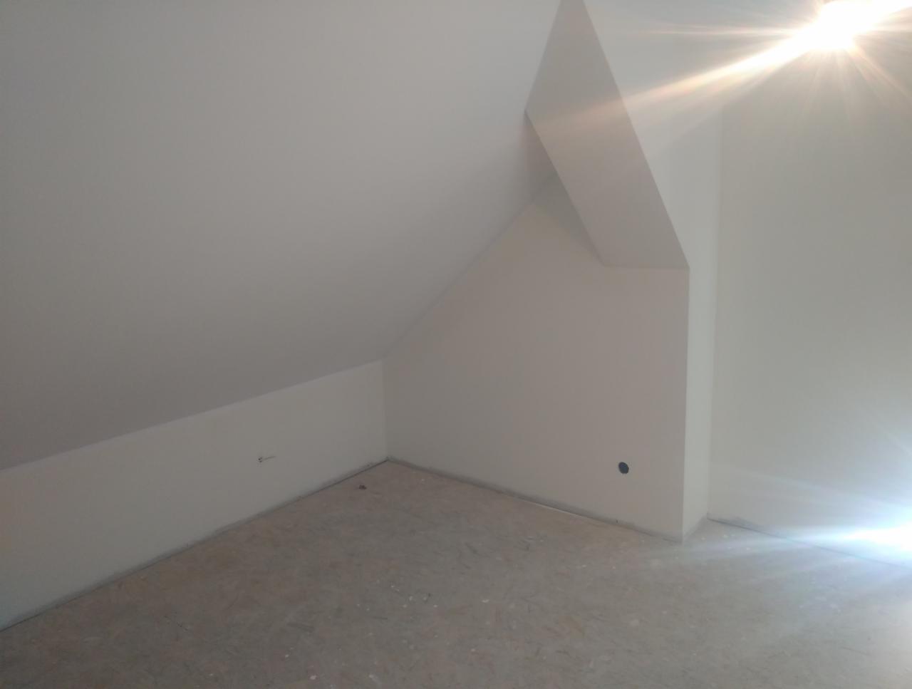 Pomalowane na biało poddasze ze skosami, widoczny surowy parkiet i ślady po gniazdku elektrycznym na ścianie.