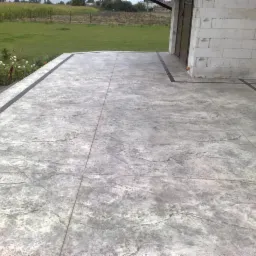 Szara, wzorzysta posadzka na zewnątrz budynku, z ciemnymi obramowaniami, widok na trawnik i pole w tle.