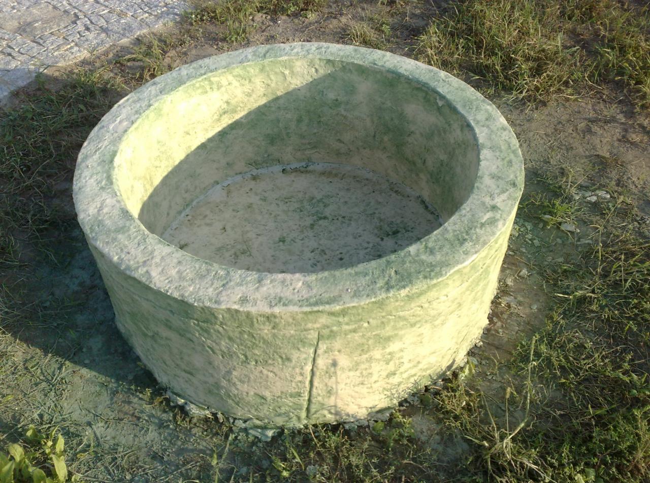 Prefabrykowany betonowy krąg studzienny o szorstkiej, zielonkawej fakturze, stojący na ziemi porośniętej trawą.