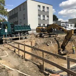 Głęboki wykop zabezpieczony drewnianą szalunką, z pracującą koparką CAT i wywrotką w tle, na tle budynku mieszkalnego w słoneczny dzień.