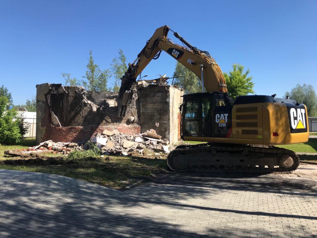 Żółta koparka CAT 329E kruszy pozostałości ceglanego budynku pod błękitnym niebem, widoczne gruz i zieleń w tle.