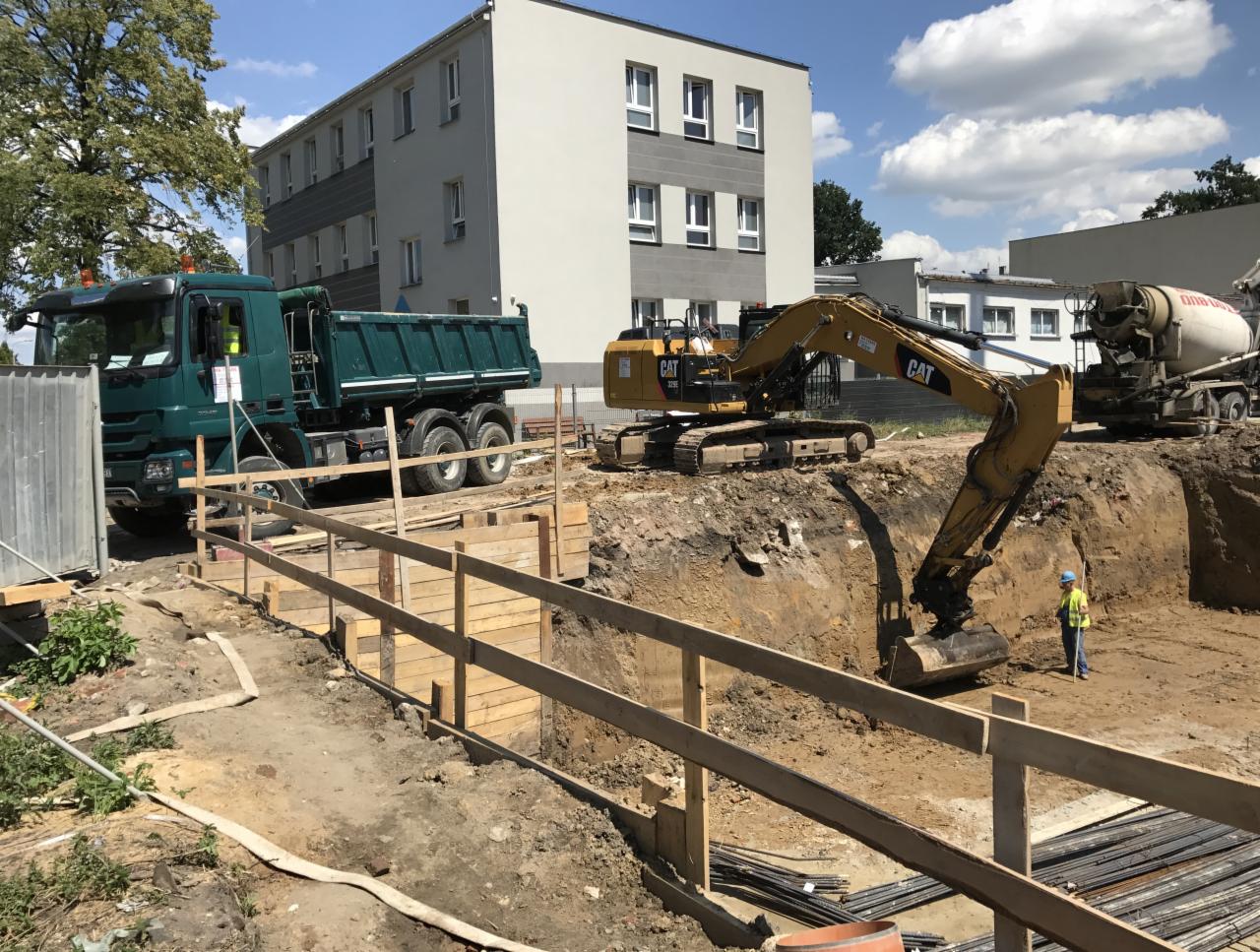 Głęboki wykop zabezpieczony drewnianą szalunką, z pracującą koparką CAT i wywrotką w tle, na tle budynku mieszkalnego w słoneczny dzień.