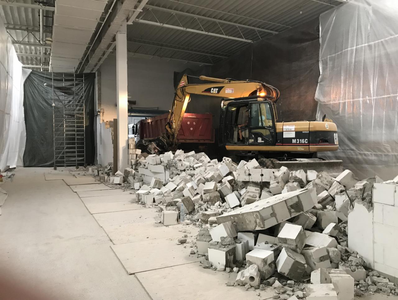 Żółta koparka CAT M316C ładuje gruz z białych bloczków do czerwonej ciężarówki wewnątrz budynku, widoczne zabezpieczenia z folii i rusztowania.