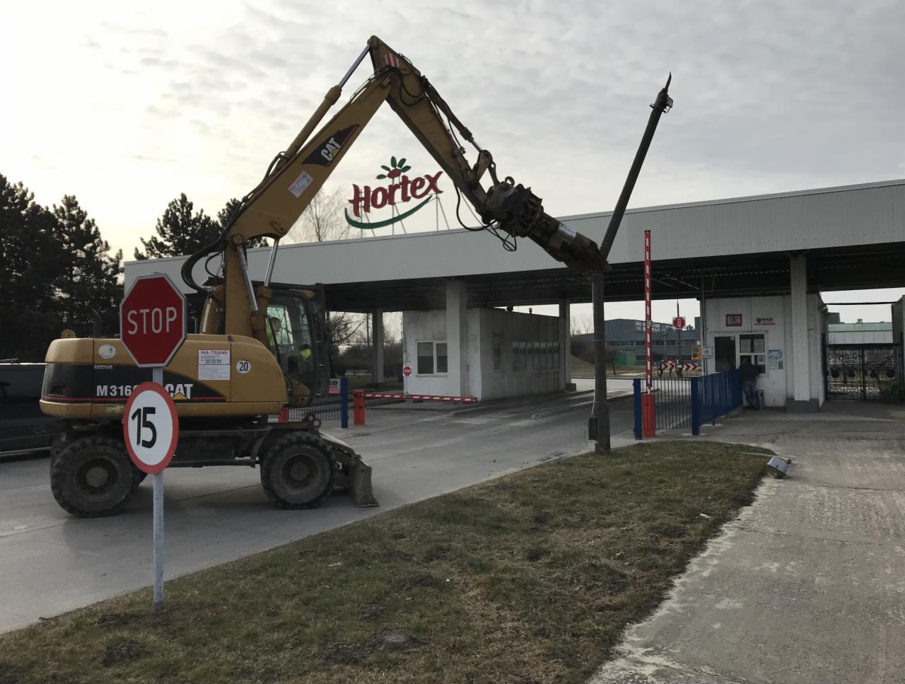 Żółta koparka CAT M316 z hydraulicznym młotem kruszącym przy bramie wjazdowej do fabryki Hortex, znak STOP i ograniczenie prędkości do 15 km/h.