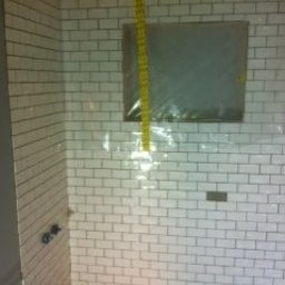 Panorenno - Wnętrze łazienki w trakcie wykańczania: białe, prostokątne płytki ułożone na ścianach, zabezpieczone okno folią, widoczne wystające rury instalacji wodnej i fragment sufitu z izolacją.