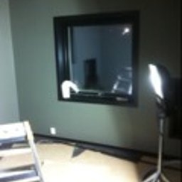 Panorenno - Wnętrze pomieszczenia w trakcie remontu: ściana pomalowana na oliwkowy kolor, czarna rama okna, drabina oparta o ścianę, lampa halogenowa na statywie oświetlająca fragment podłogi zabezpieczonej...