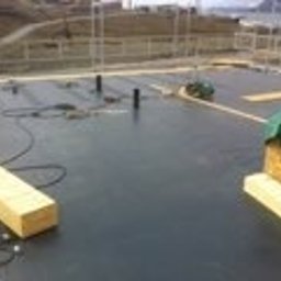 Panorenno - Czarna, płaska powierzchnia balkonu w trakcie prac izolacyjnych, widoczne elementy konstrukcyjne, materiały izolacyjne przykryte zieloną folią, kable i drewniane elementy. W tle widoczne budynki...