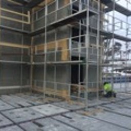 Panorenno - Budynek w trakcie ocieplania, widoczne rusztowania, płyty izolacyjne i fragmenty drewnianej konstrukcji. Prace prowadzone na wysokości, w tle krajobraz miejski.