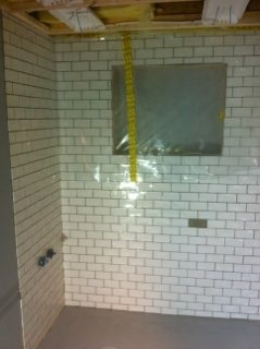 Wnętrze łazienki w trakcie wykańczania: białe, prostokątne płytki ułożone na ścianach, zabezpieczone okno folią, widoczne wystające rury instalacji wodnej i fragment sufitu z izolacją.