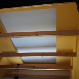 Sufit z żółtymi ścianami i drewnianymi belkami, widok z dołu, w tle okno i roślina doniczkowa.