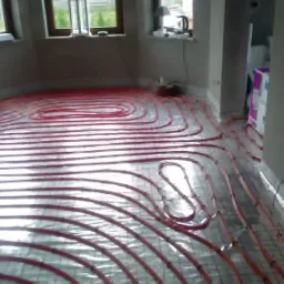 Czerwone rury ogrzewania podłogowego ułożone na folii odblaskowej w surowym pomieszczeniu z oknami.