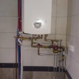 Biały wiszący piec gazowy Termet z podłączeniami rur miedzianych i stalowych na tle kafelkowanej ściany, widoczny fragment grzejnika i kratki wentylacyjnej.