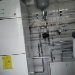 Biała jednostka pompy ciepła z widocznymi rurami instalacji grzewczej, zbiornik wyrównawczy i żółta naklejka ostrzegawcza na tle kafelkowanej ściany.