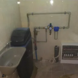 Wnętrze pomieszczenia z zainstalowanym zmiękczaczem wody obok szarej umywalki, rury instalacji wodnej z filtrami i manometrem na ścianie z beżowych płytek oraz biała szafka rozdzielcza ogrzewania...