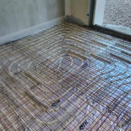 Widok z góry na rozłożoną instalację ogrzewania podłogowego z rurami w metalowej siatce, ułożoną na folii izolacyjnej w surowym wnętrzu z widocznym progiem drzwi balkonowych i posadzką na zewnątrz.