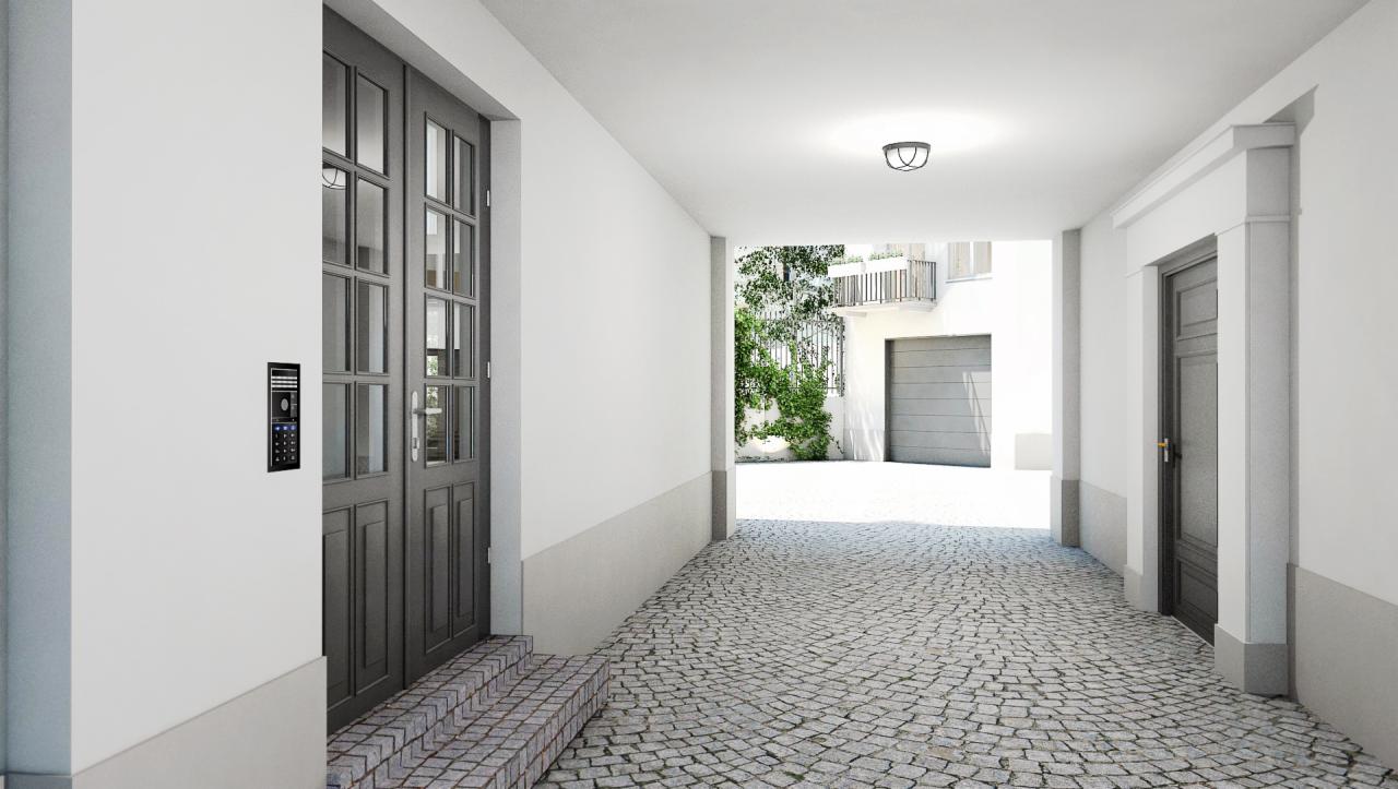 Wejście do budynku z szarymi drzwiami, domofonem i brukowaną ścieżką prowadzącą do garażu w tle, oświetlone światłem dziennym.