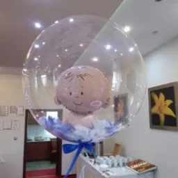 Przezroczysty balon wypełniony mniejszym balonem w kształcie głowy niemowlęcia z niebieskimi piórkami i kokardą, ustawiony w pomieszczeniu z czerwonym dywanem i stołem z przekąskami.