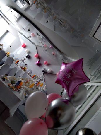 Uroczystość w sali z dekoracjami balonowymi: różowe, srebrne i białe balony, w tym jeden w kształcie gwiazdy z napisem, oraz nakryte stoły z jedzeniem w tle.