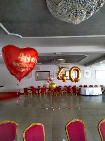 Dekoracja sali na 40. rocznicę ślubu z czerwonym balonem w kształcie serca i złotymi balonami cyframi, widoczne krzesła i stół z białym obrusem, oświetlenie sufitowe.