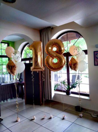 Dekoracja sali na 18 urodziny: złote balony foliowe w kształcie cyfr '1' i '8' oraz bukiety balonów z helem w kolorach białym, złotym i transparentnym z konfetti, ustawione na podłodze w jasnym...