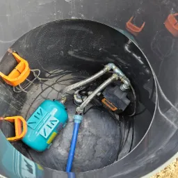 Wnętrze czarnej studni z widocznym osprzętem: hydrofor w kolorze turkusowym, czarna pompa, rury ze stali nierdzewnej, niebieski wąż oraz uchwyty z tworzywa w kolorze pomarańczowym.