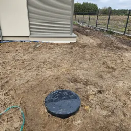 Czarna pokrywa studzienki rewizyjnej w ziemi obok budynku z szarą roletą okienną i fragmentem niebieskiego węża, widoczny ogrodzenie z siatki i drzewa w tle.