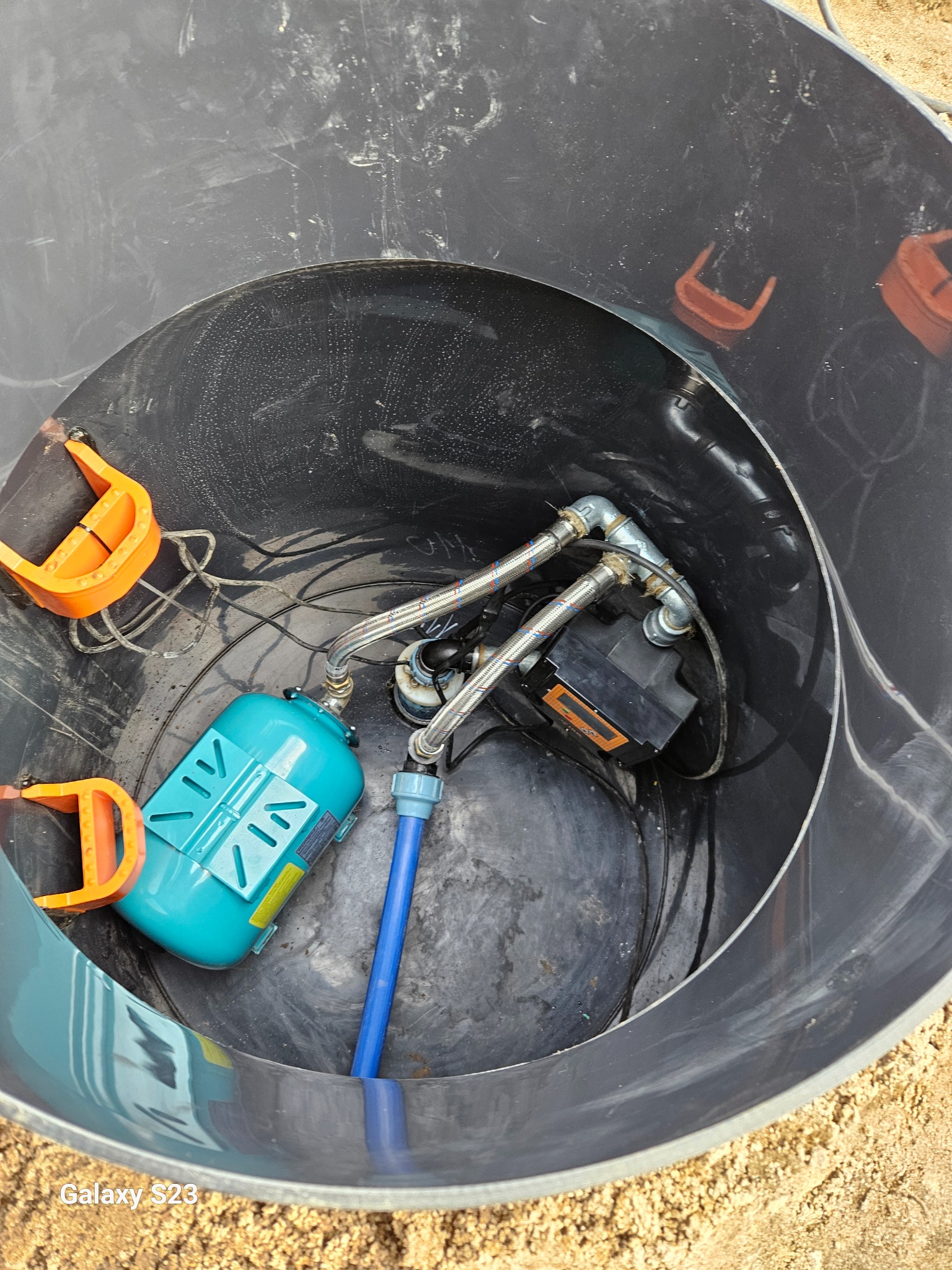 Wnętrze czarnej studni z widocznym osprzętem: hydrofor w kolorze turkusowym, czarna pompa, rury ze stali nierdzewnej, niebieski wąż oraz uchwyty z tworzywa w kolorze pomarańczowym.