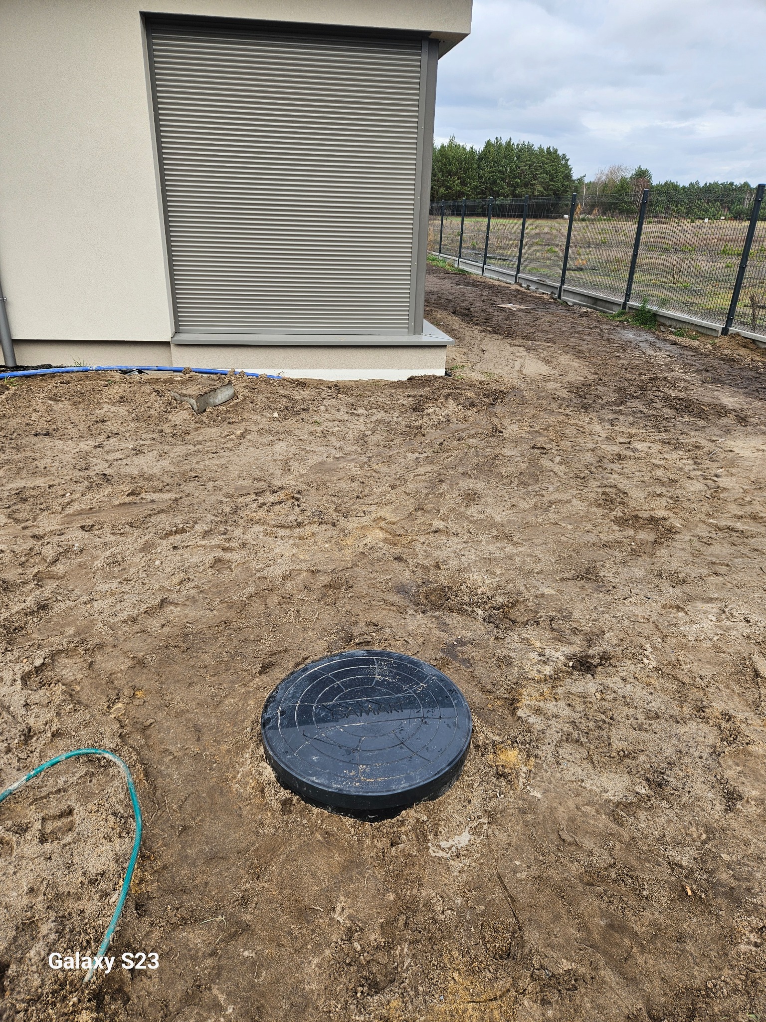 Czarna pokrywa studzienki rewizyjnej w ziemi obok budynku z szarą roletą okienną i fragmentem niebieskiego węża, widoczny ogrodzenie z siatki i drzewa w tle.