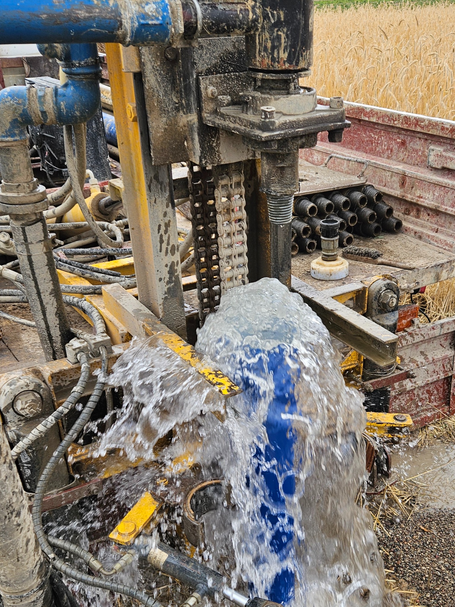 Woda tryskająca z maszyny wiertniczej na tle pola zboża, widoczne elementy hydrauliczne i rury.