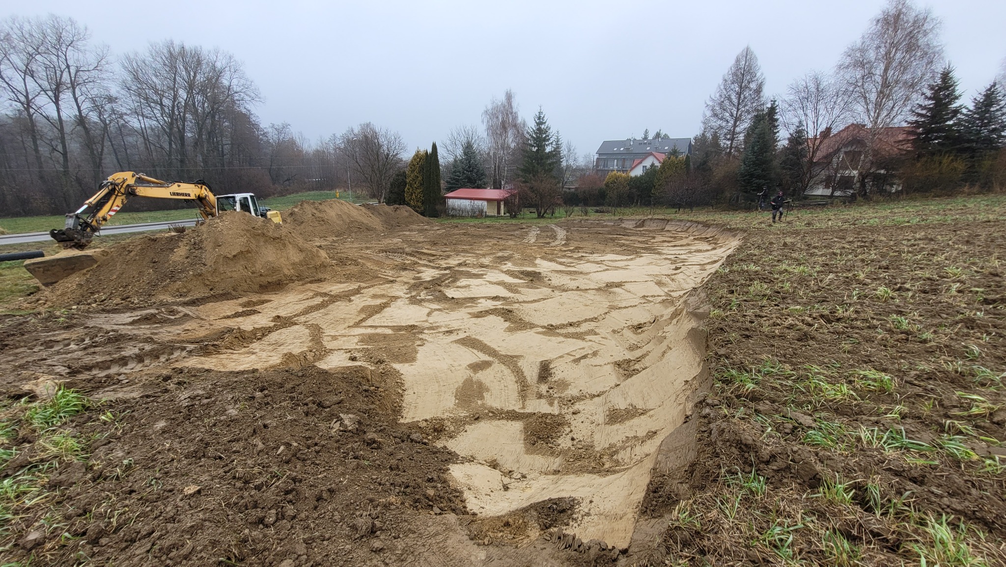 Żółta koparka Liebherr przy wykopie ziemnym na działce, widoczne ślady opon, w tle zabudowania mieszkalne i drzewa pod pochmurnym niebem.