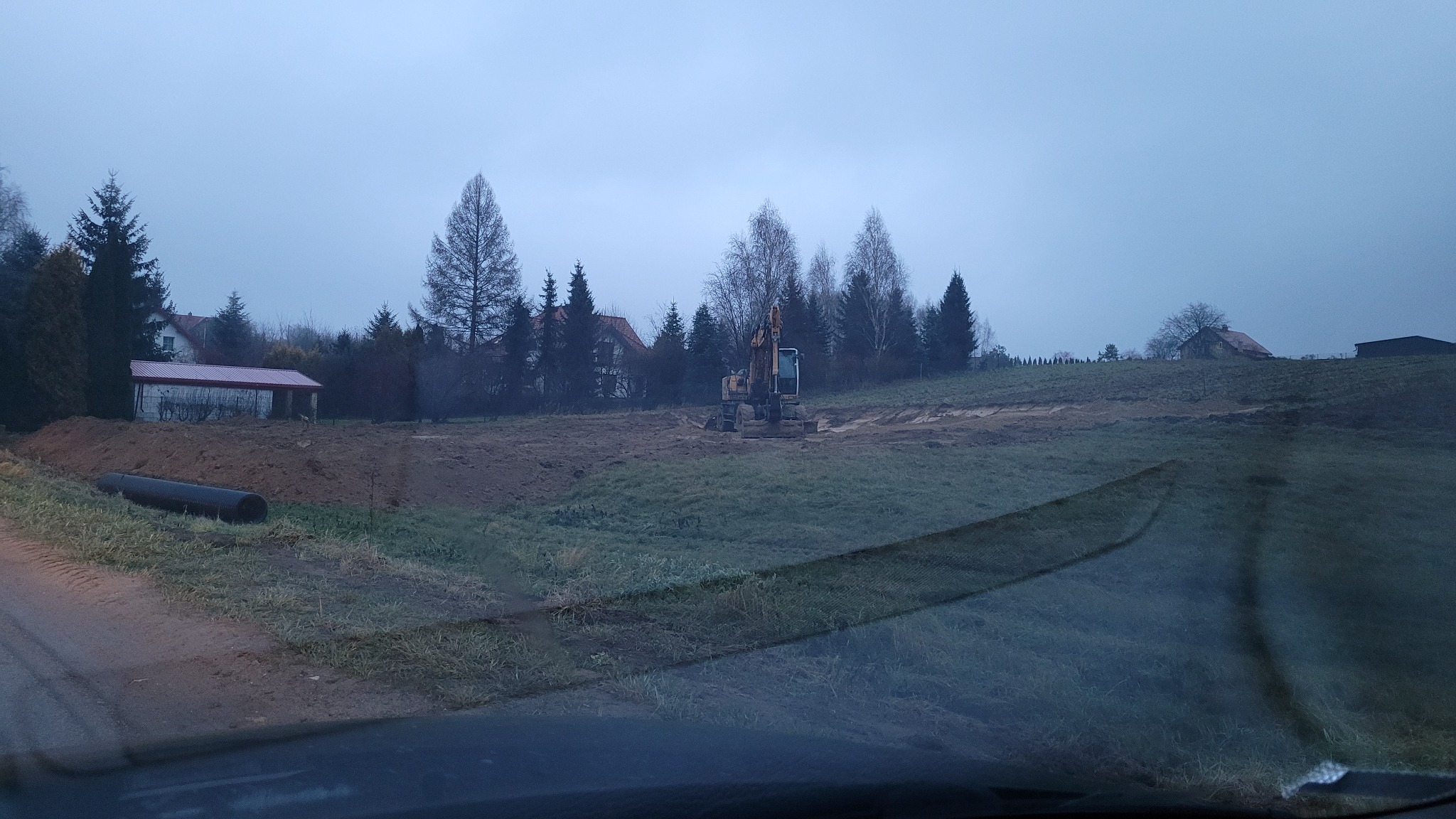 Żółta koparka na placu budowy, widoczna rura kanalizacyjna, w tle budynki mieszkalne i drzewa w pochmurny dzień.
