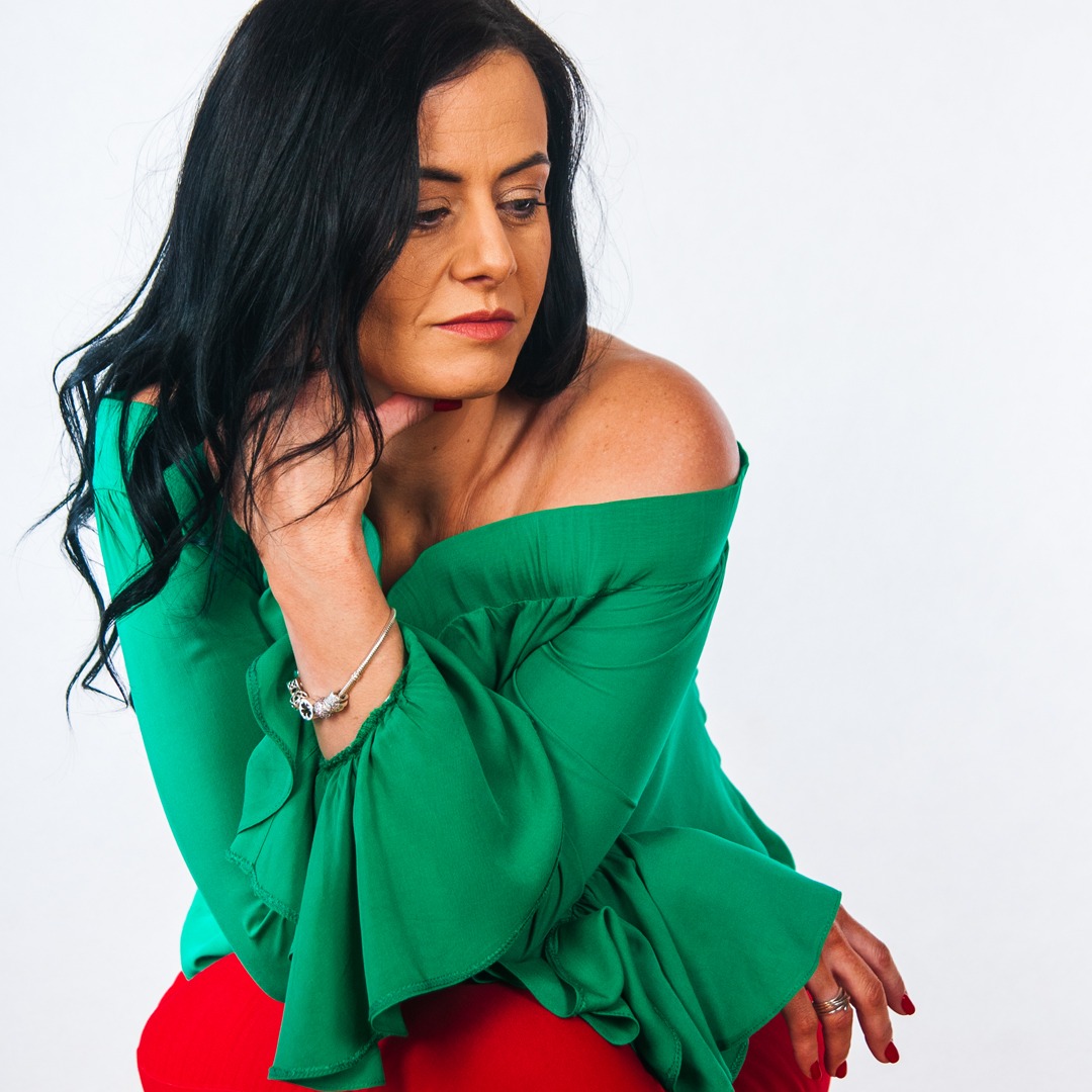 Portret kobiety z ciemnymi włosami, ubrana w zieloną bluzkę z odkrytymi ramionami i czerwone spodnie, pozująca na białym tle, zamyślony wyraz twarzy.