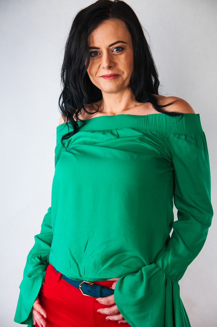 Portret kobiety w zielonej bluzce z odkrytymi ramionami i czerwonych spodniach, pozującej na jasnym tle.