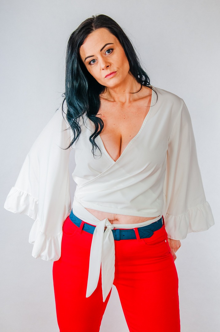 Kobieta w białej bluzce z falbanami i czerwonych spodniach, pozująca na jasnym tle, z ręką schowaną za plecami.