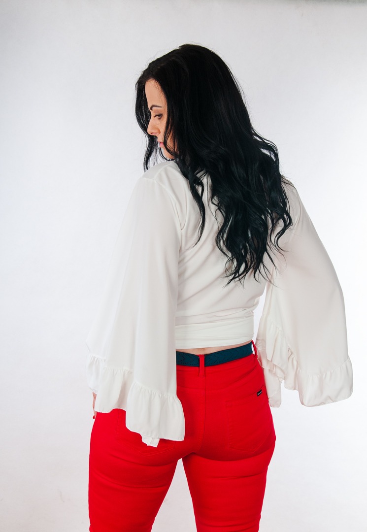 Kobieta w białej bluzce z szerokimi rękawami i czerwonych spodniach, odwrócona tyłem, na jasnym tle.